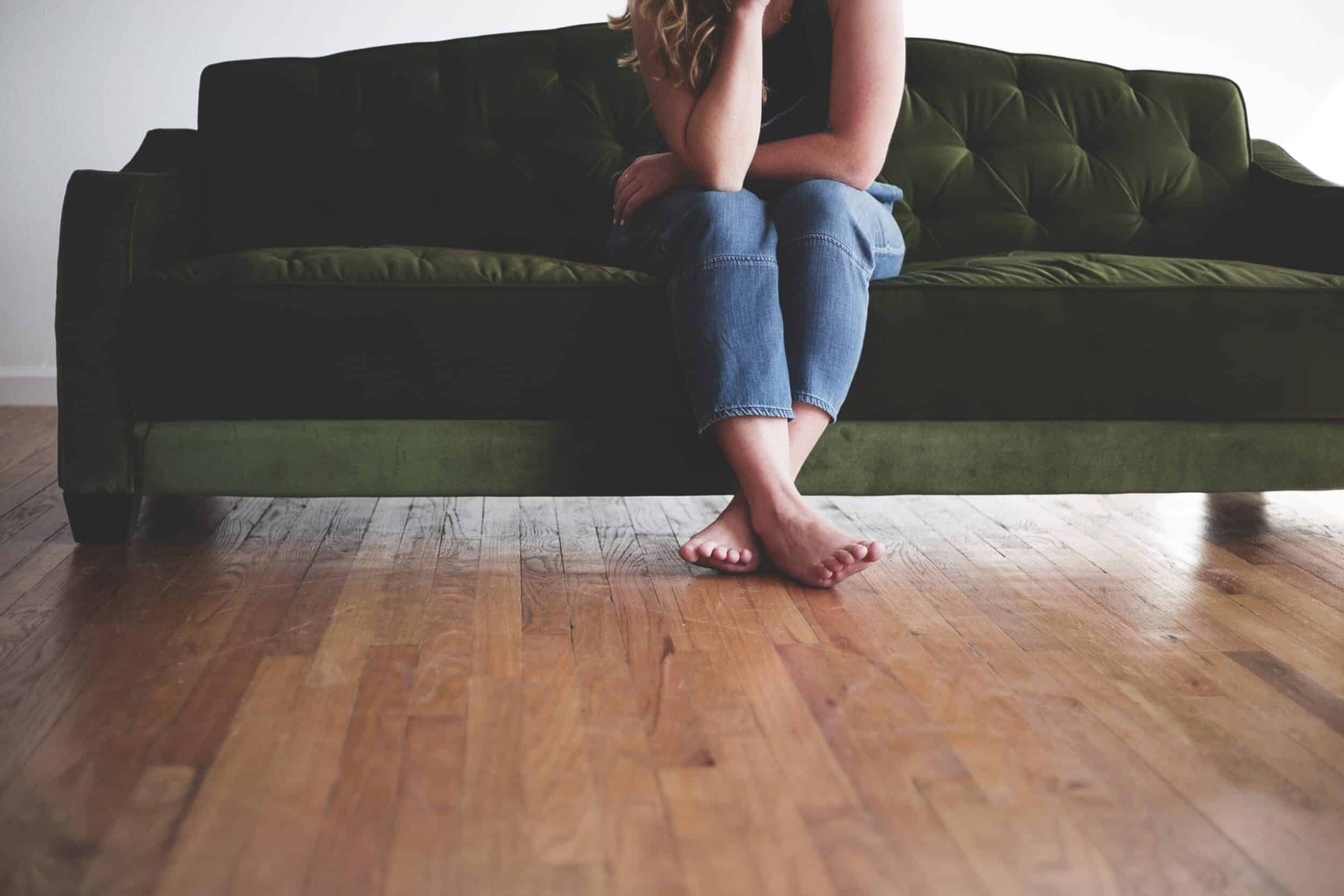 Horizontal shot of a barefoot woman in blue jeans sitting on a green couch deep in thought